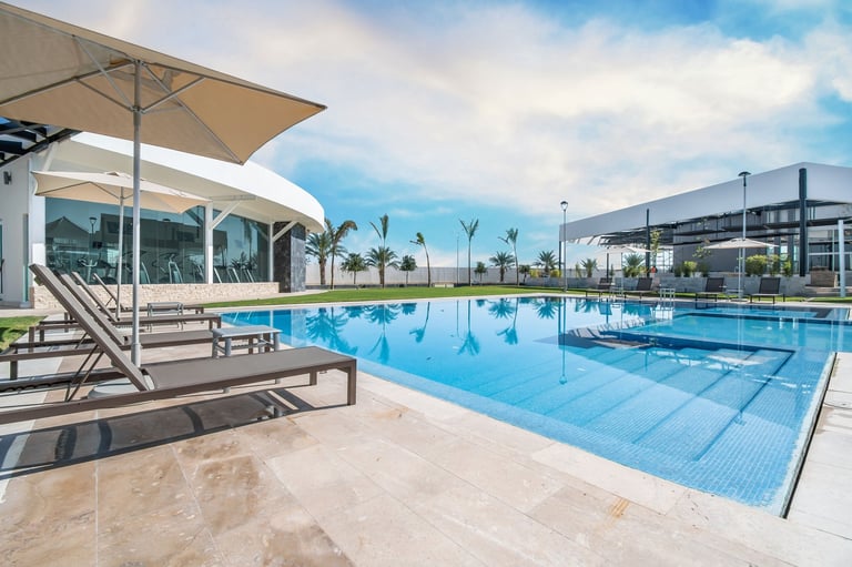Modern swimming pool facility with clear blue water, sun loungers under umbrellas, and glass building overlooking the pool deck