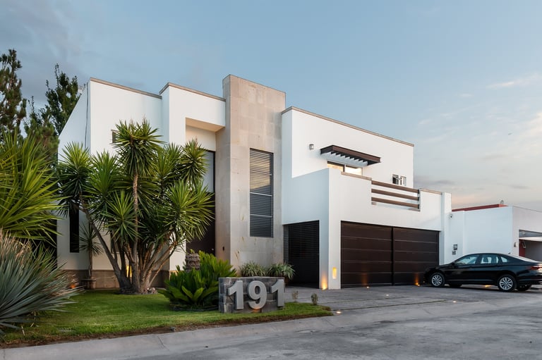 Modern white house with geometric architecture, palm trees, and dark garage doors. Address number 191 visible on the lawn.