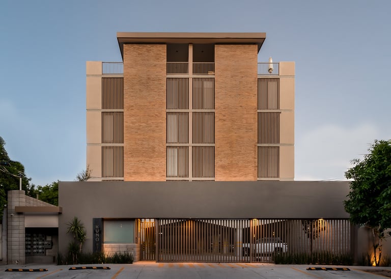 Modern brick residential building with geometric facade, gold-toned metal gates, and landscaping at dusk