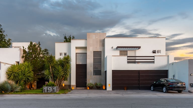 Modern minimalist house with white and tan exterior, dark garage doors, driveway with black car, and palm trees at dusk