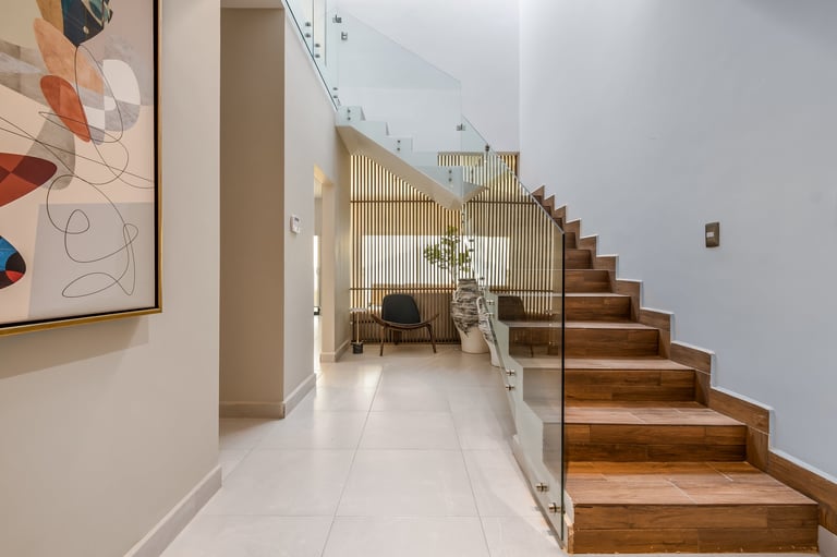 Modern hallway with wooden staircase on right, white tile flooring, and mezzanine office area with glass partition and workspace nook above stairs